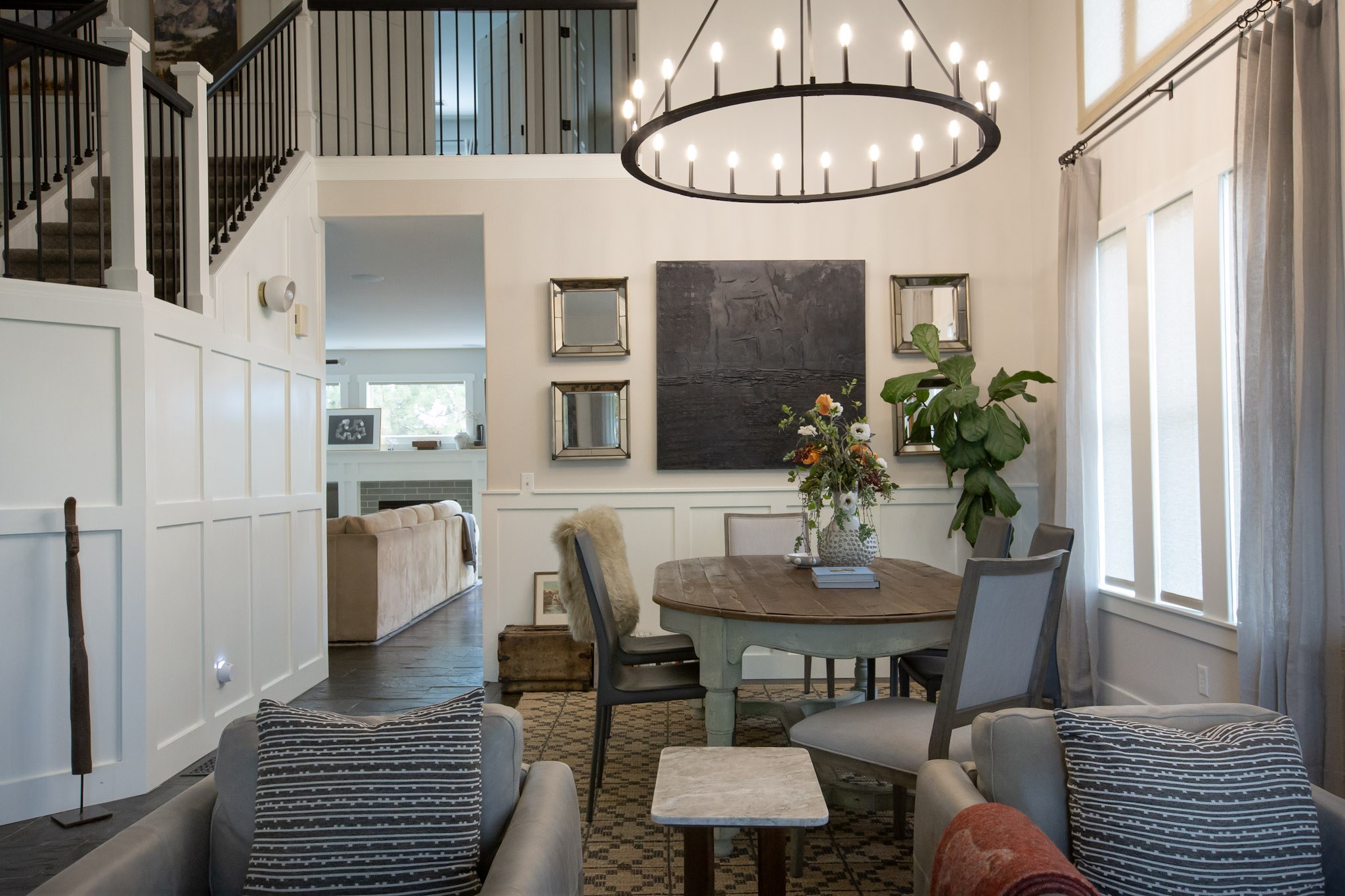 A bright dining area with a round farmhouse table, a large iron ring chandelier, paired mirrors, and a fiddle-leaf fig by the window