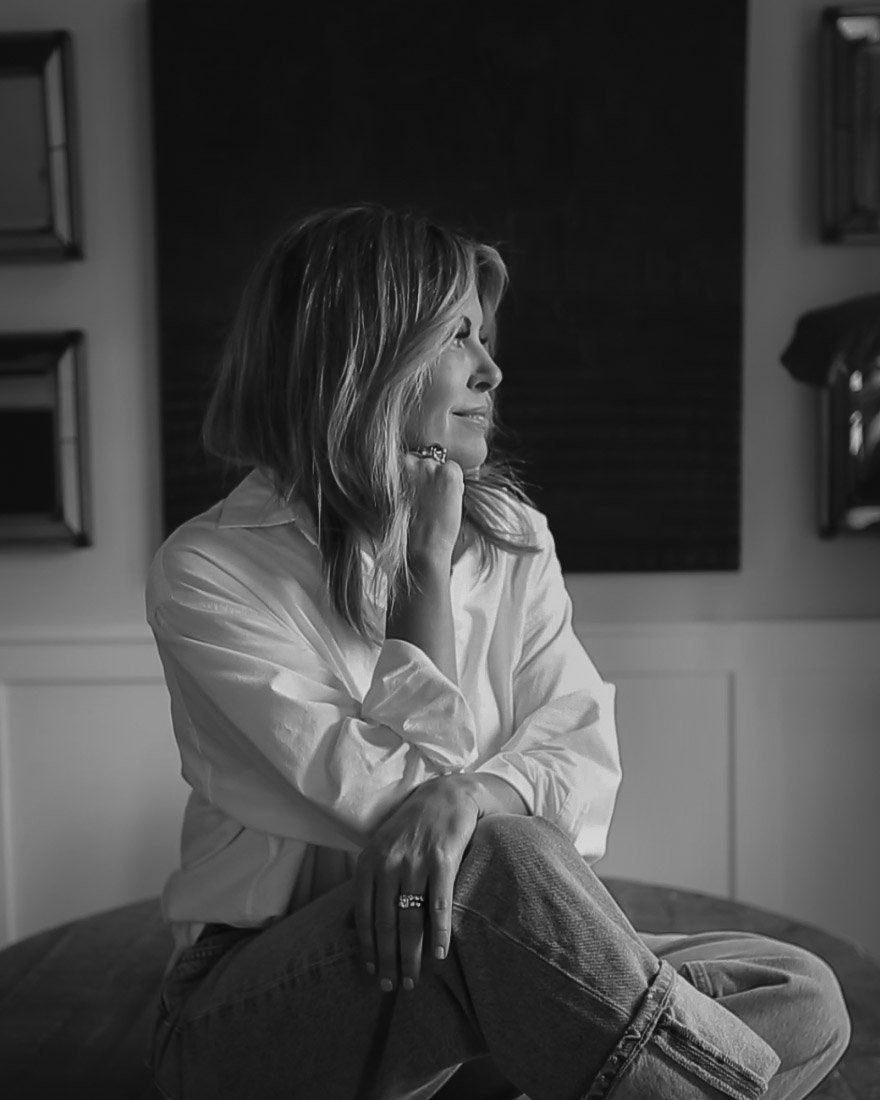 Robyn, designer and founder of Everyday Grace Collective, sitting on a table in a white shirt and jeans, chin resting on her hand, looking toward the window