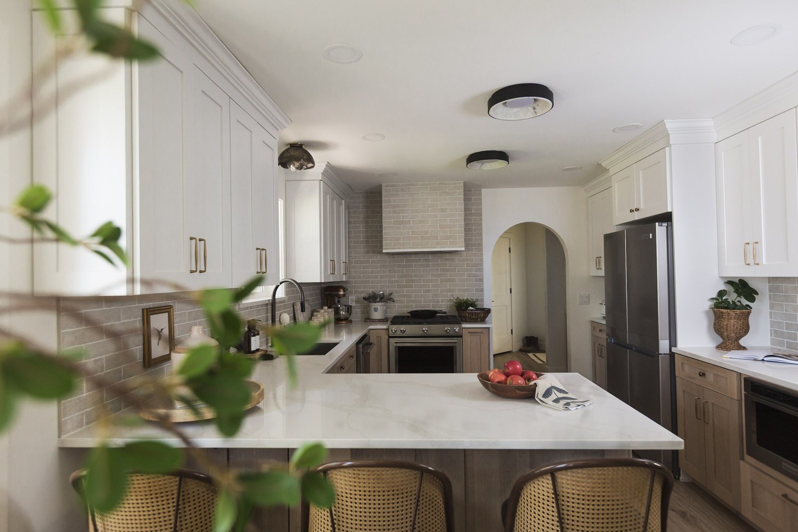 A white and oak kitchen in soft daylight, with a marble peninsula, cane counter stools, gray tile backsplash, and an arched doorway beyond