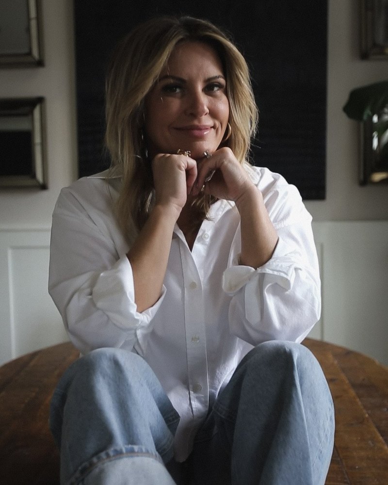 Robyn, designer and founder of Everyday Grace Collective, seated on a wooden table in a white shirt and jeans, chin resting on her hands