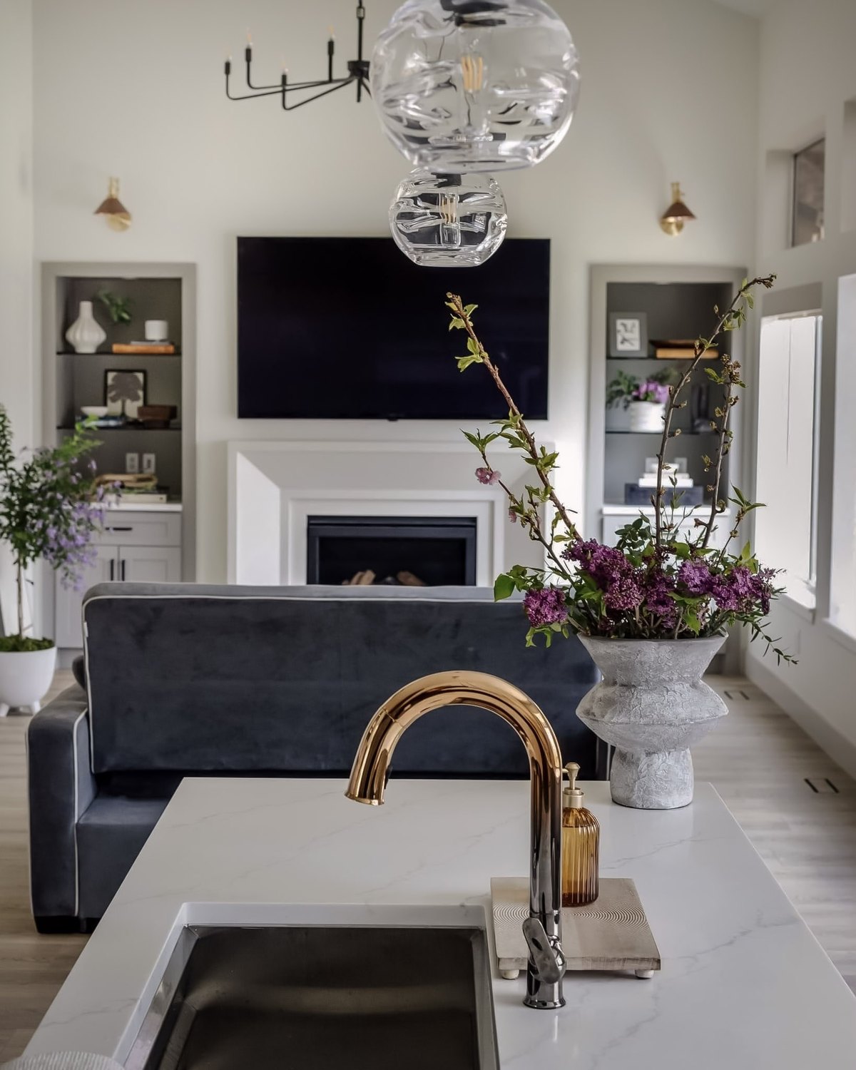 A living room seen across a kitchen island, with lilacs in a stone vase, a navy velvet sofa, and a fireplace beyond