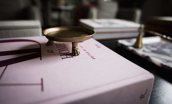 A small footed brass dish resting on a pale pink design book, with more books and a marble tray in soft low light