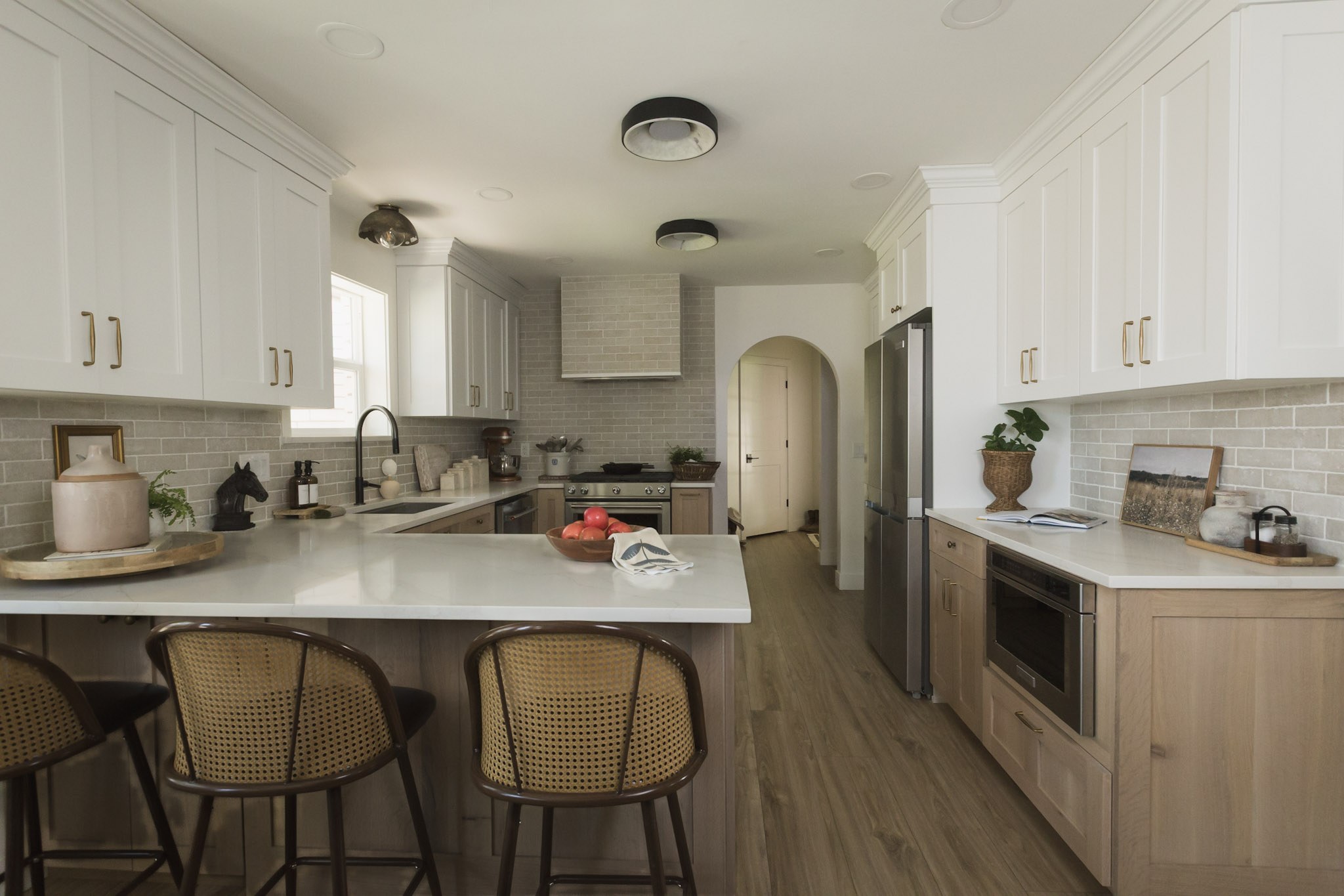 A finished white and oak kitchen with a marble peninsula, cane counter stools, gray tile backsplash, and an arched doorway