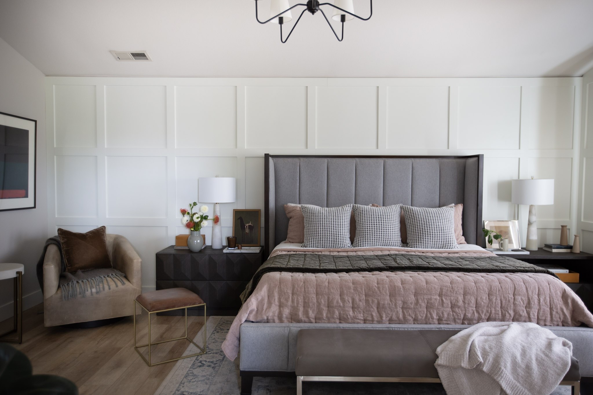 A primary bedroom with a tall gray upholstered headboard, dusty pink quilt, houndstooth pillows, and a paneled accent wall