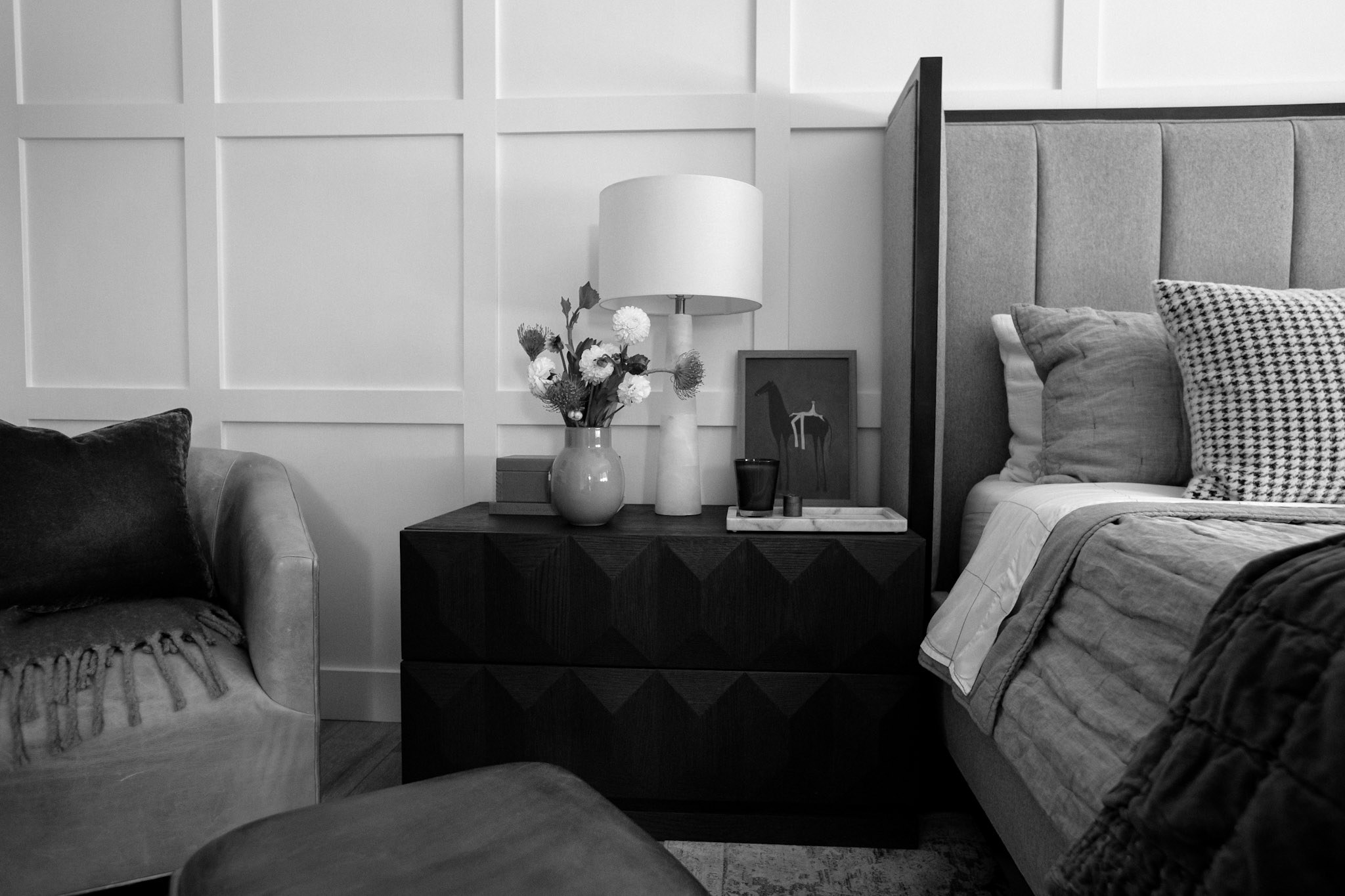 A black and white view of the bedroom, showing the nightstand, upholstered bed, and a velvet armchair