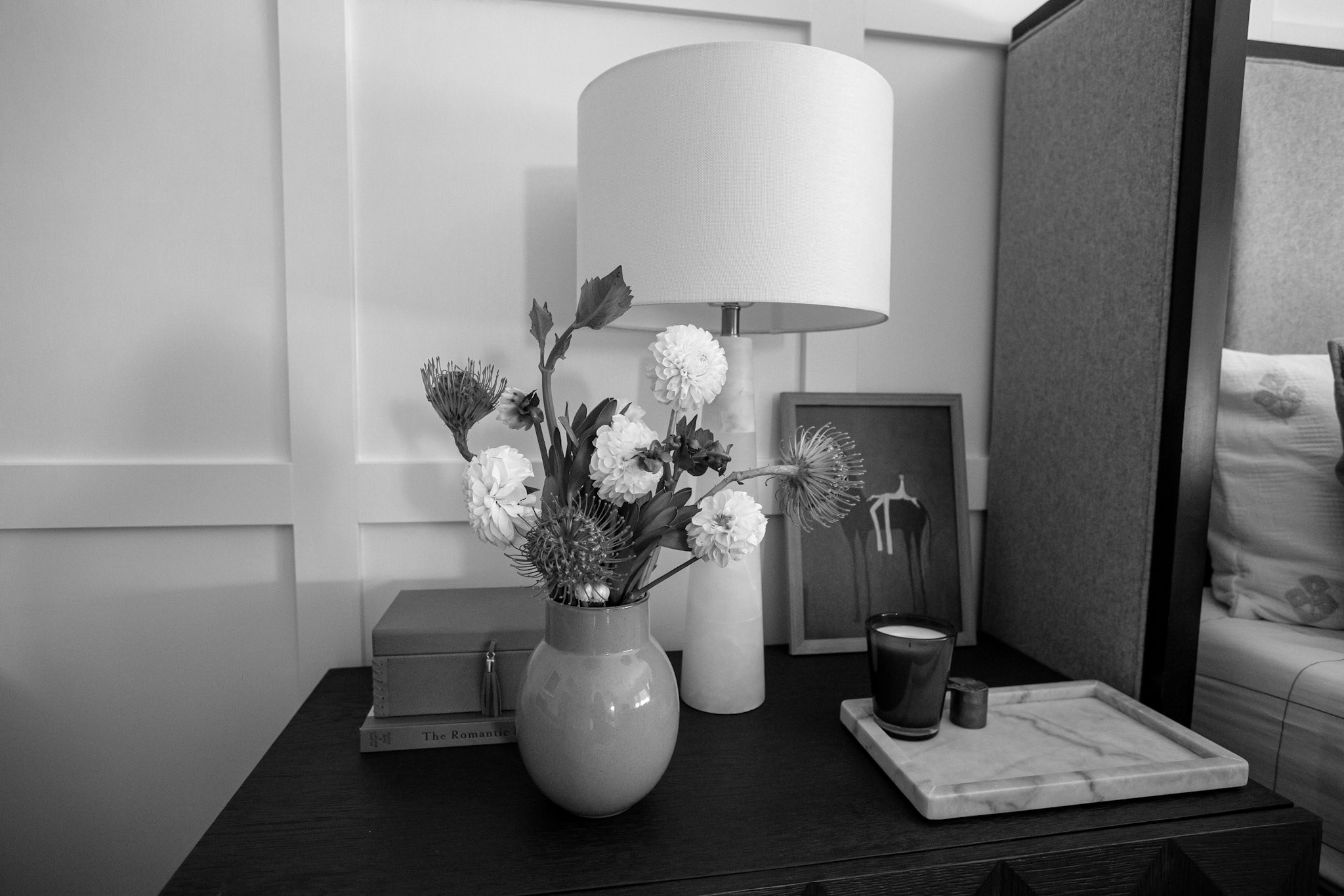 A black and white still life of the bedside table with an alabaster lamp, dahlias in a vase, and a candle on a marble tray