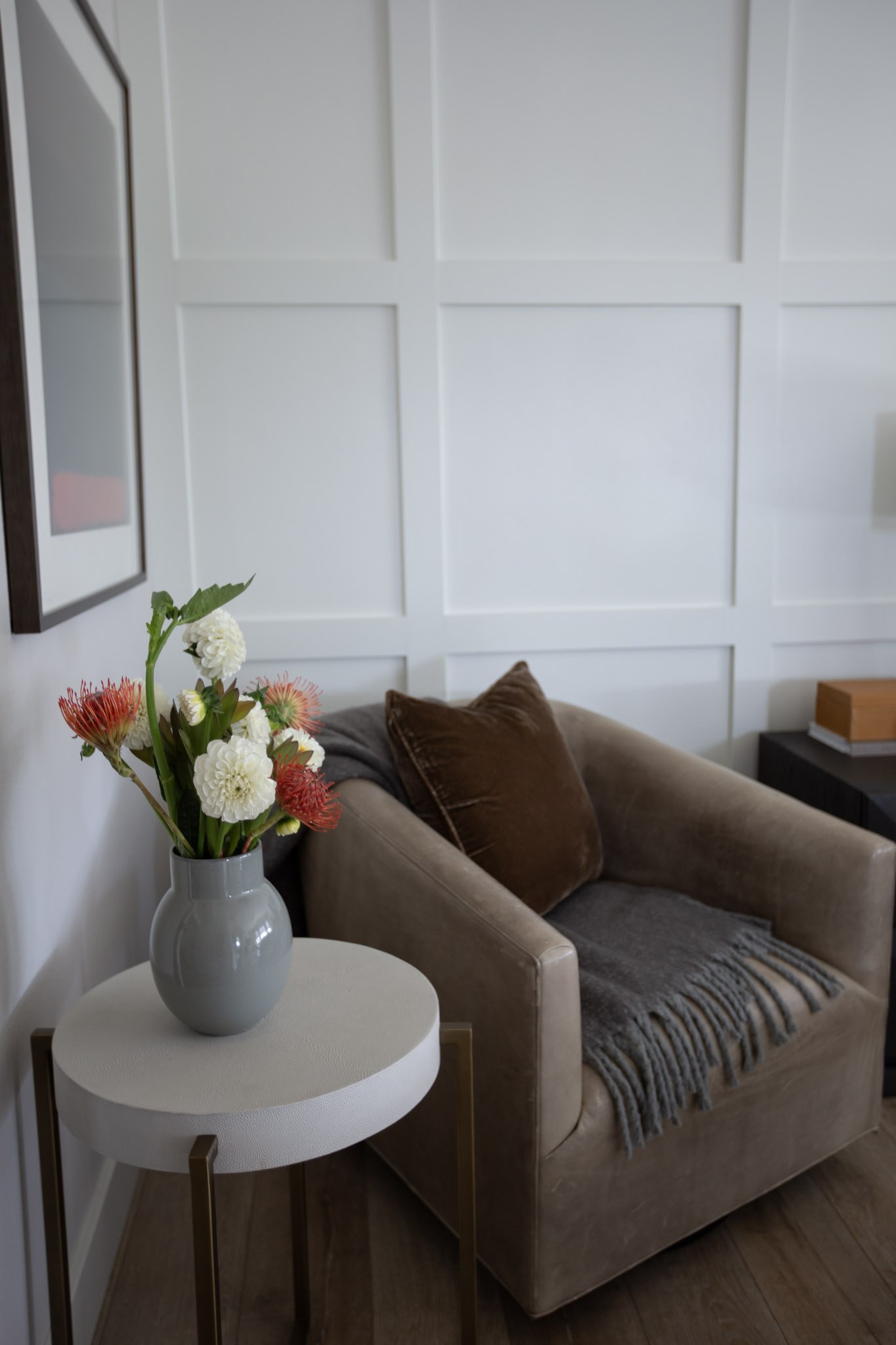 A leather club chair with a velvet pillow and wool throw, beside a round side table holding dahlias in a gray vase