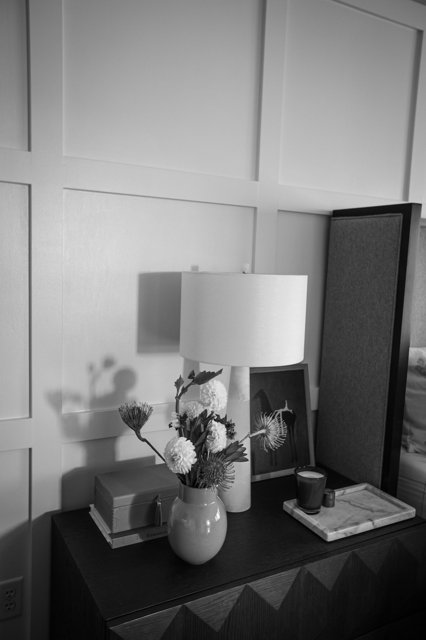 A black and white view of the faceted nightstand, lamp, and flowers against the paneled wall