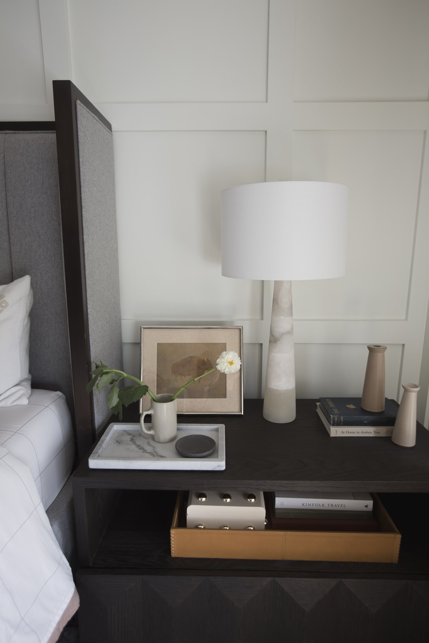 A dark wood nightstand beside the bed, styled with an alabaster lamp, a marble tray, and a single bloom in a ceramic mug