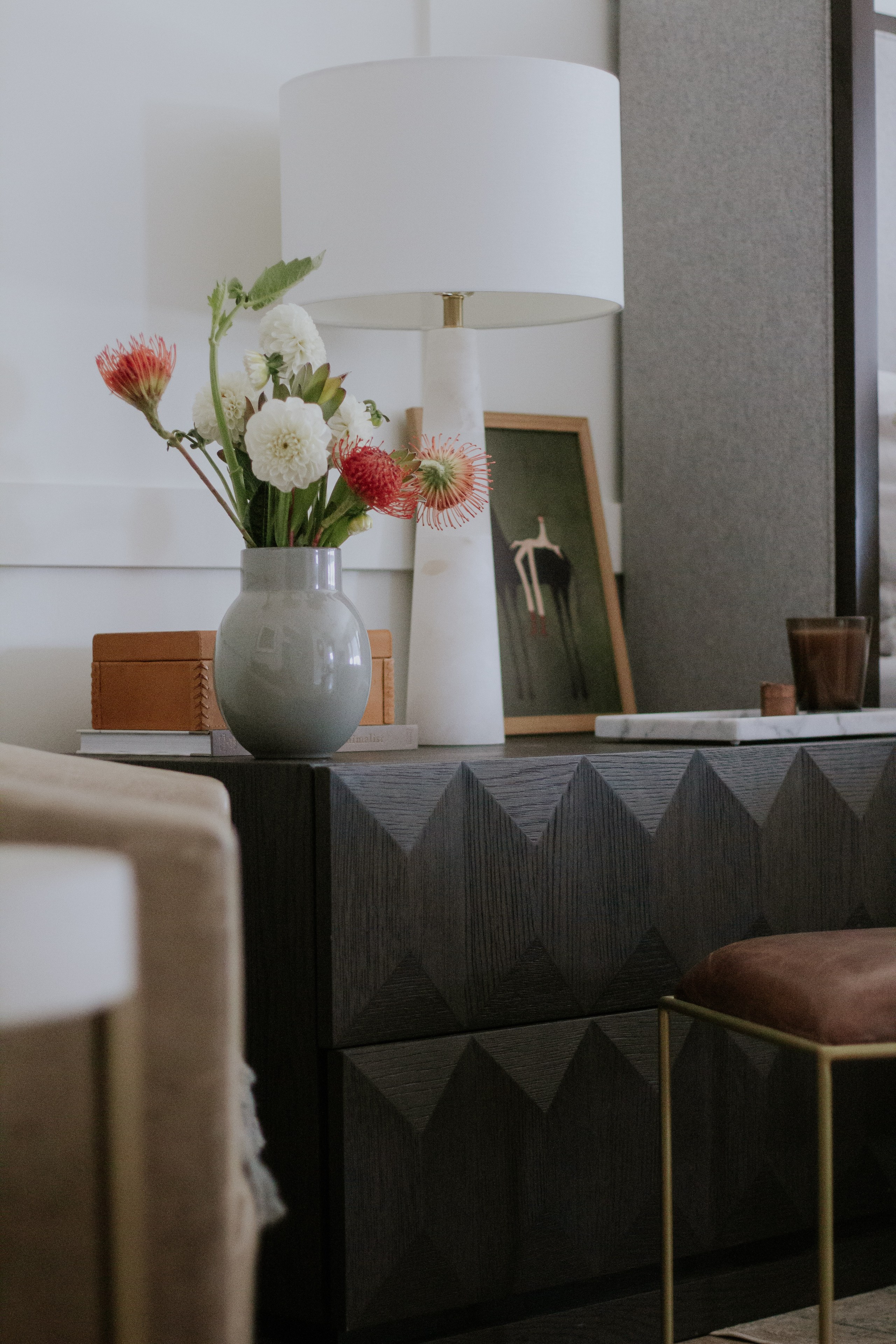 A dark faceted credenza styled with an alabaster lamp, a leather box, and white dahlias with red pincushion protea