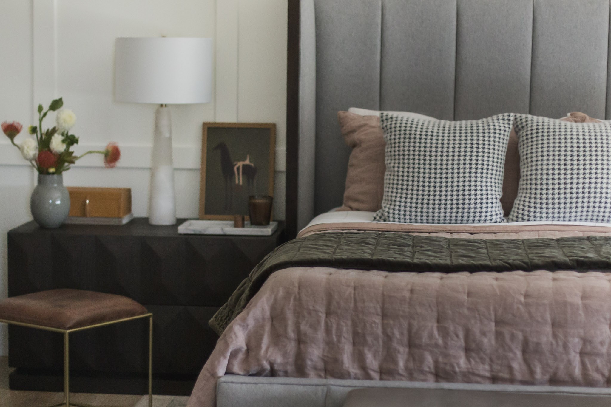 The bedroom from the foot of the bed, with layered pink and olive bedding and a brass stool beside the nightstand