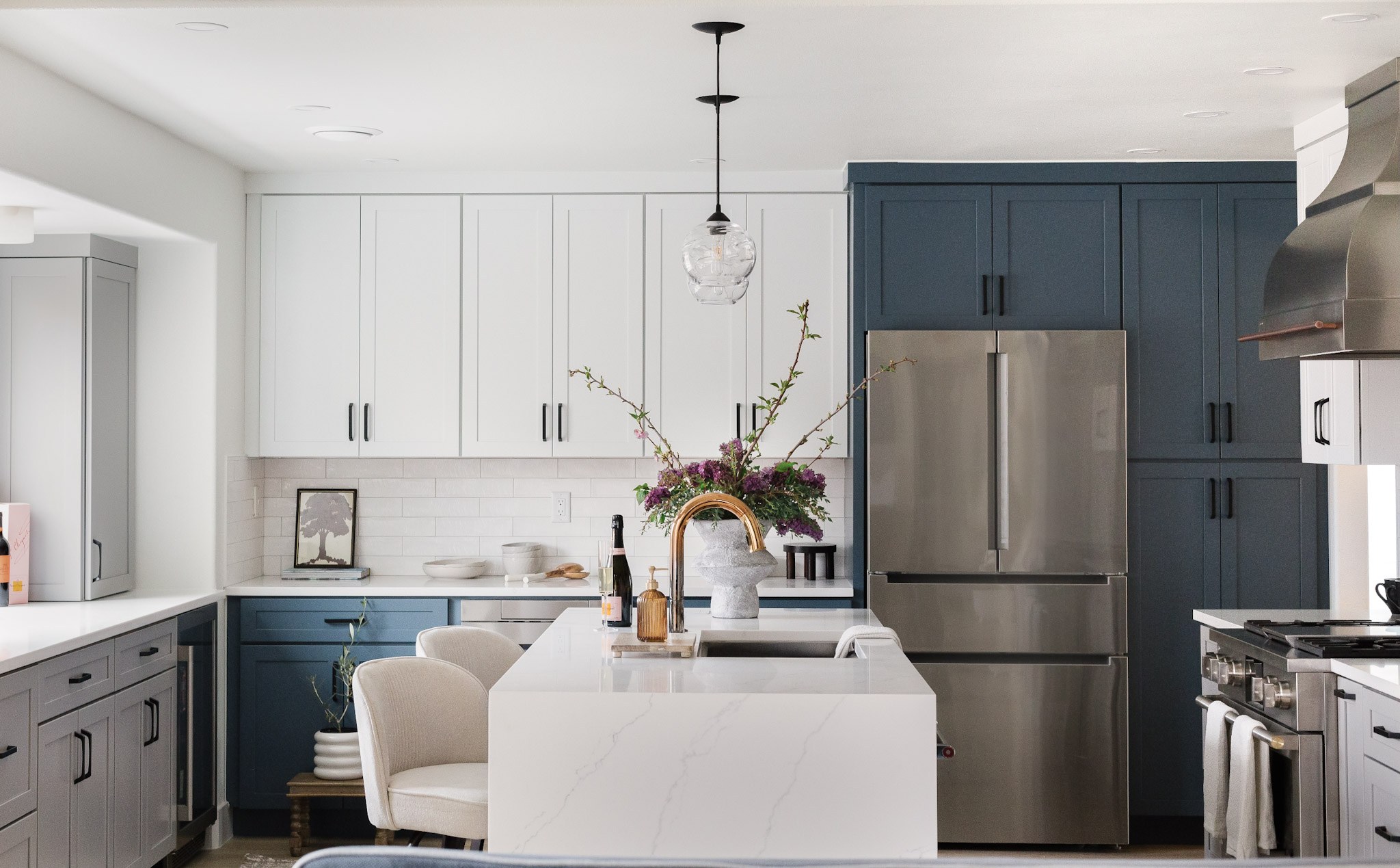 A kitchen with white upper cabinets, deep blue lower and pantry cabinetry, a waterfall quartz island, and lilacs in a stone vase