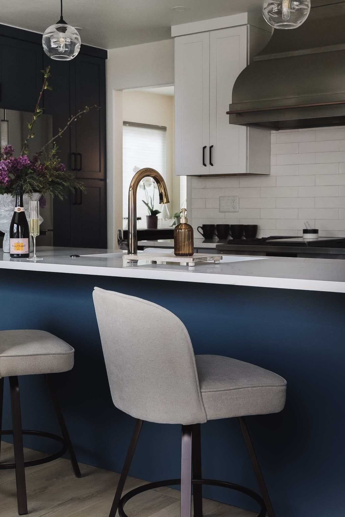 A brass faucet on a white quartz counter above a navy island, with upholstered stools and a bottle of champagne