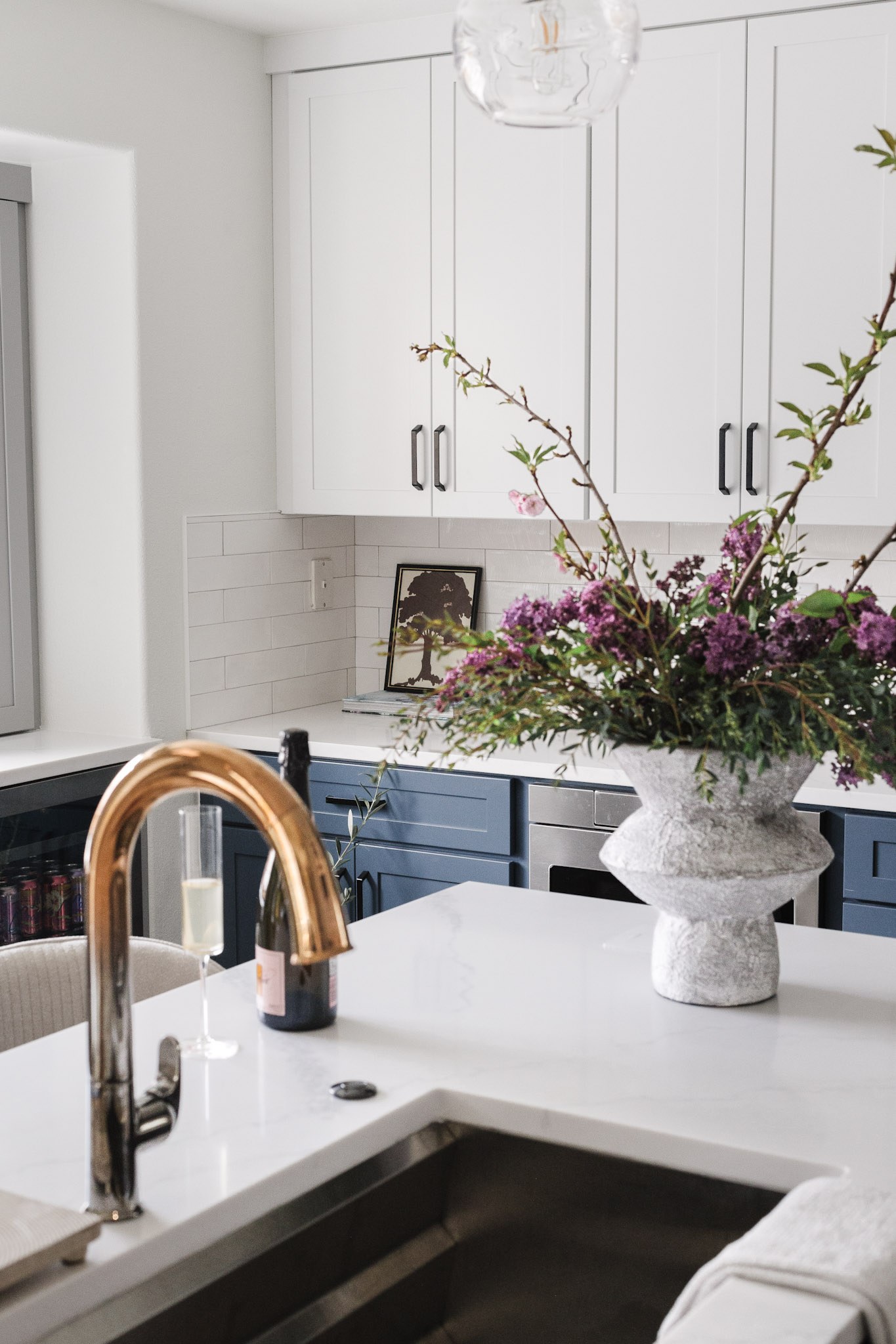 Lilacs and blossoming branches in a rough stone vase on the island, with blue cabinetry and a framed tree print behind