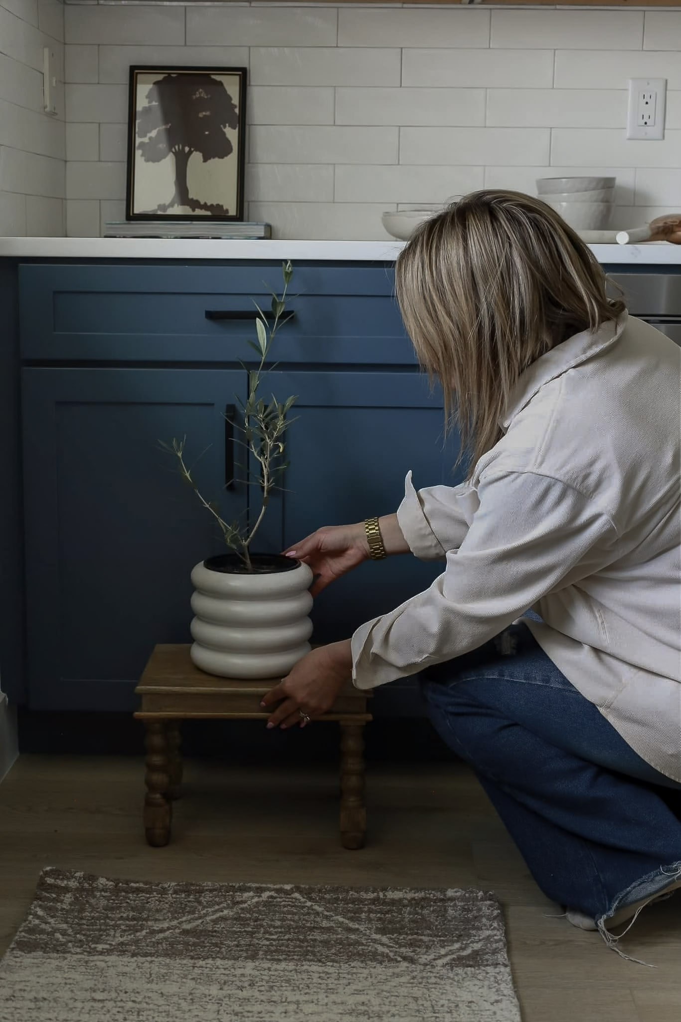 A designer settling a small olive tree in a ribbed ceramic pot onto a wooden stool beside the blue cabinetry