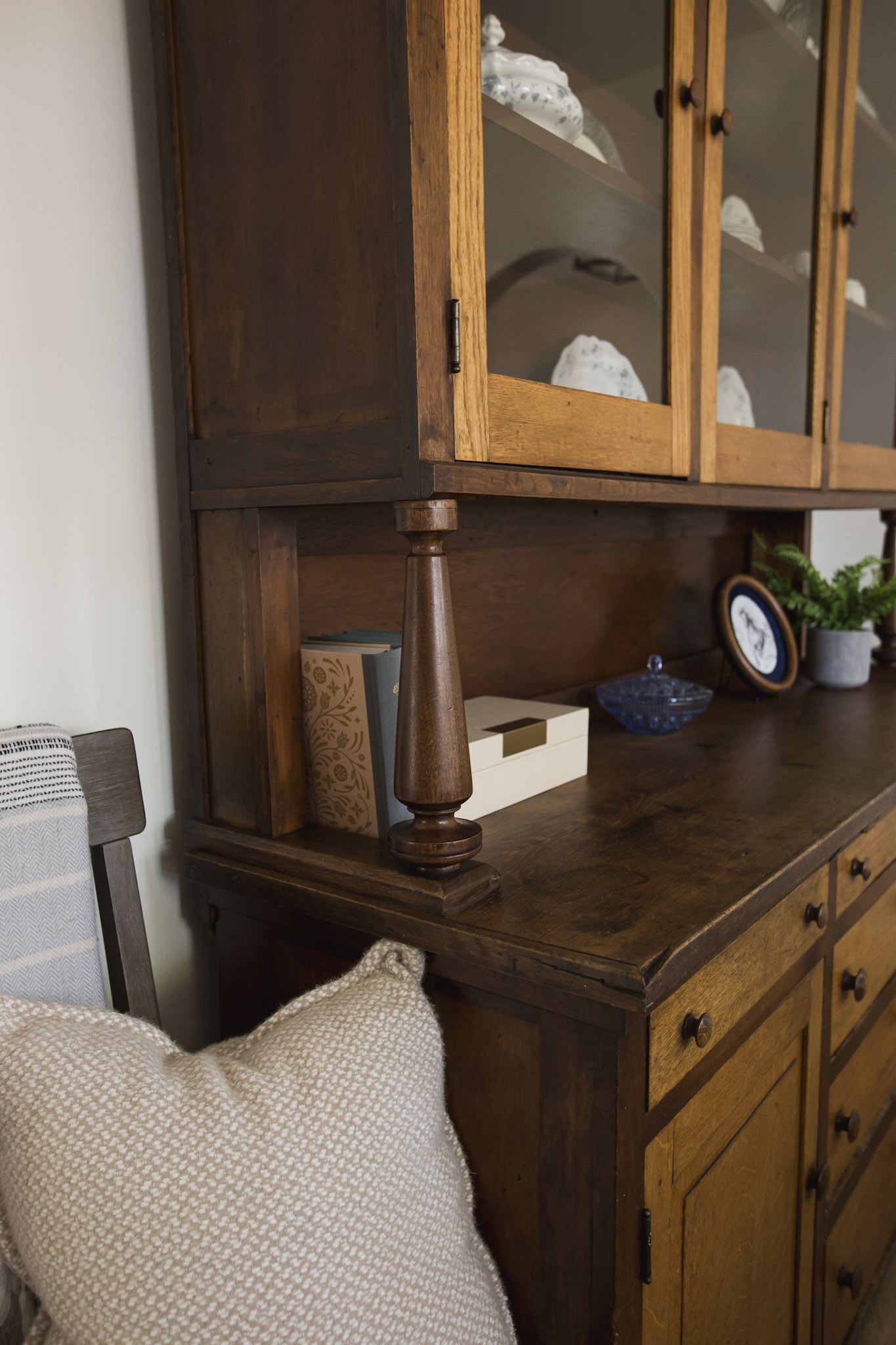 An antique wood hutch holding white ironstone behind glass doors, with books and a woven pillow in the foreground