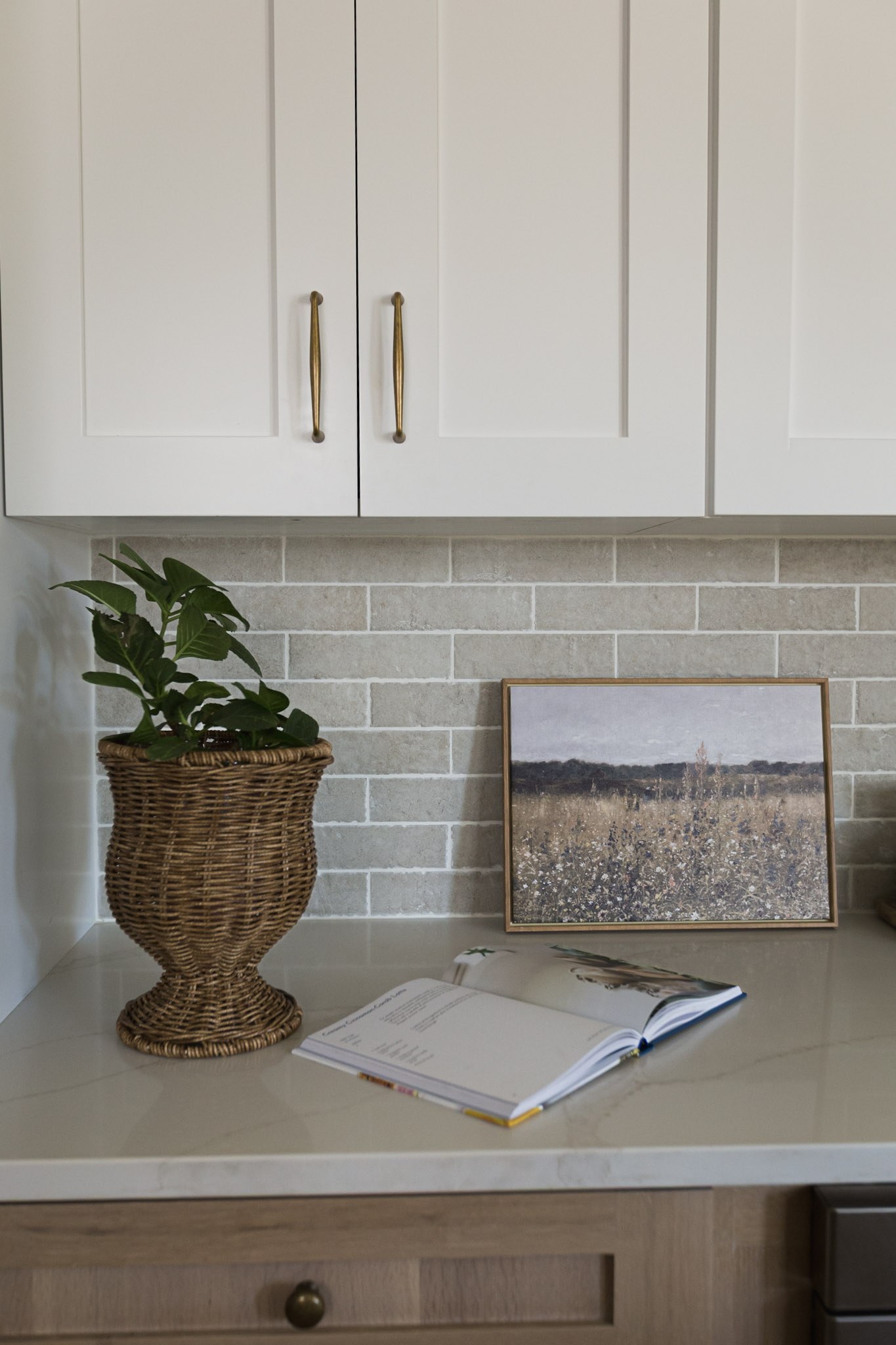 A counter corner with a wicker urn planter, a framed meadow painting, and an open cookbook
