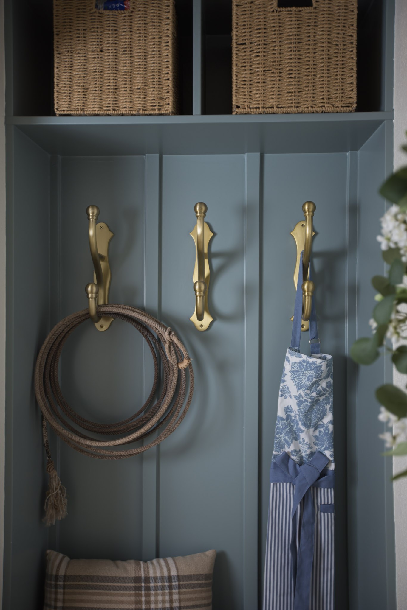 Brass hooks on a blue-green paneled wall holding a coiled rope and a blue floral apron, with woven baskets above