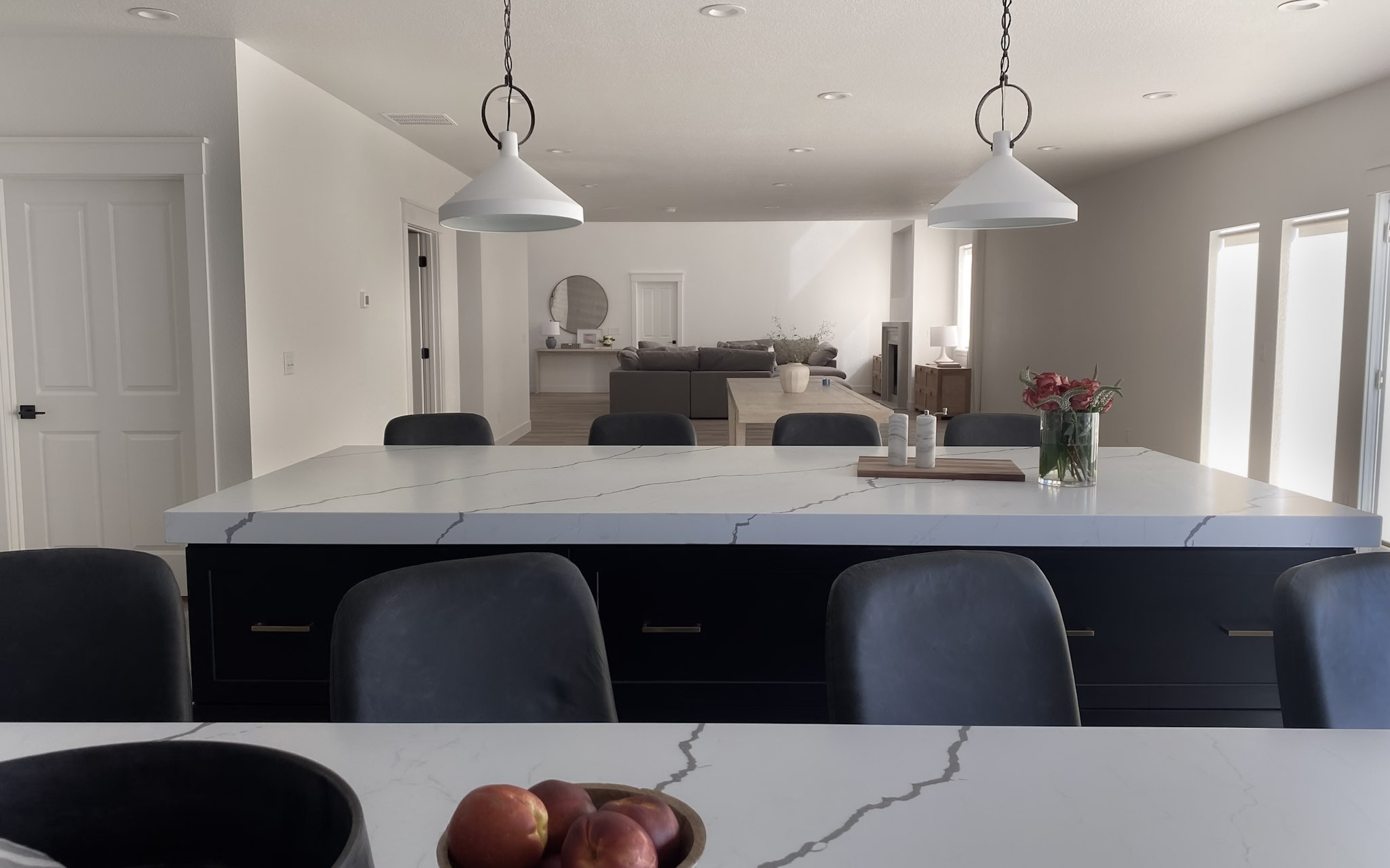 A long black kitchen island under white cone pendants, topped in veined white quartz and open to the living room beyond