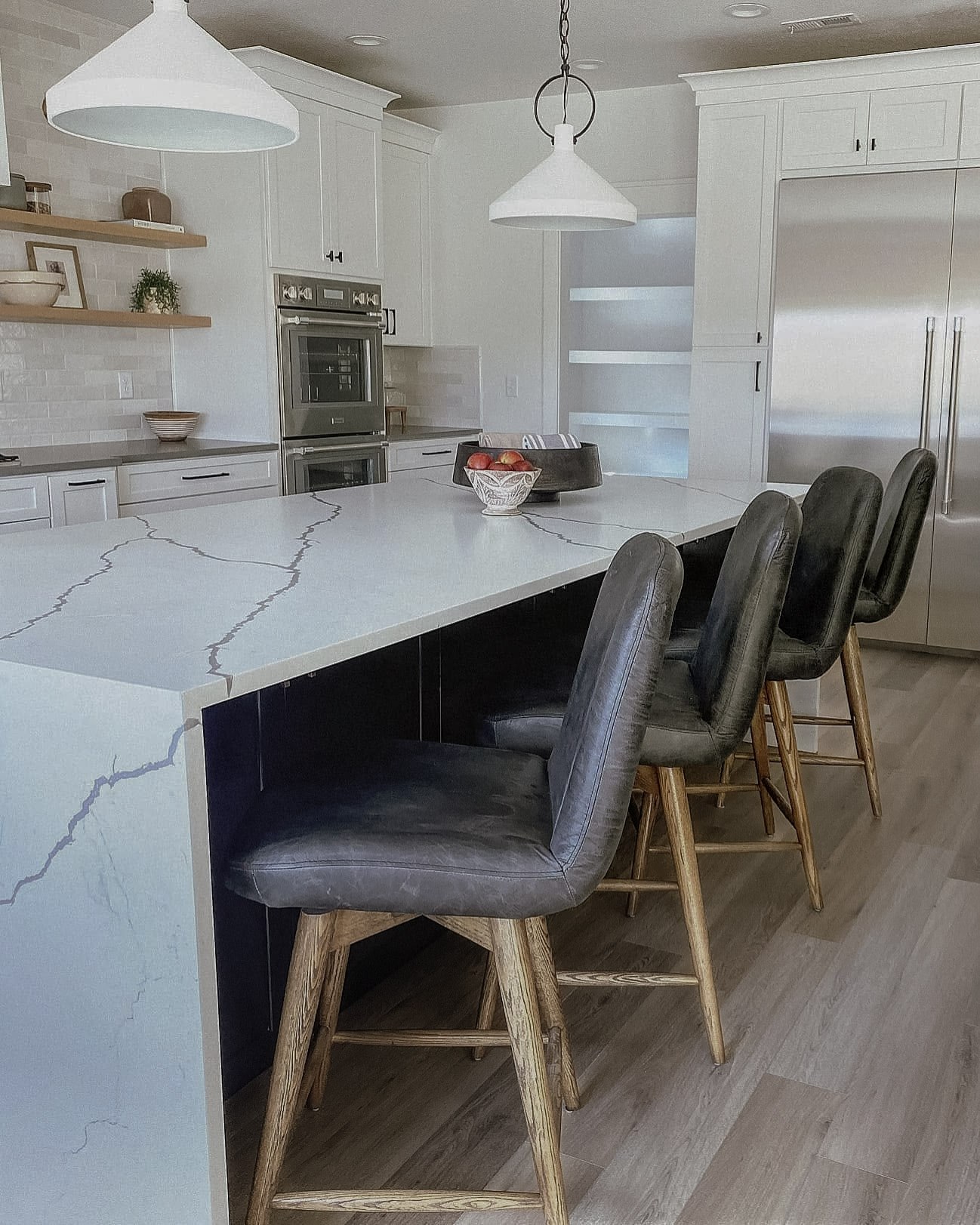 The island from the side, with dark leather counter stools, double wall ovens, and open oak shelving behind