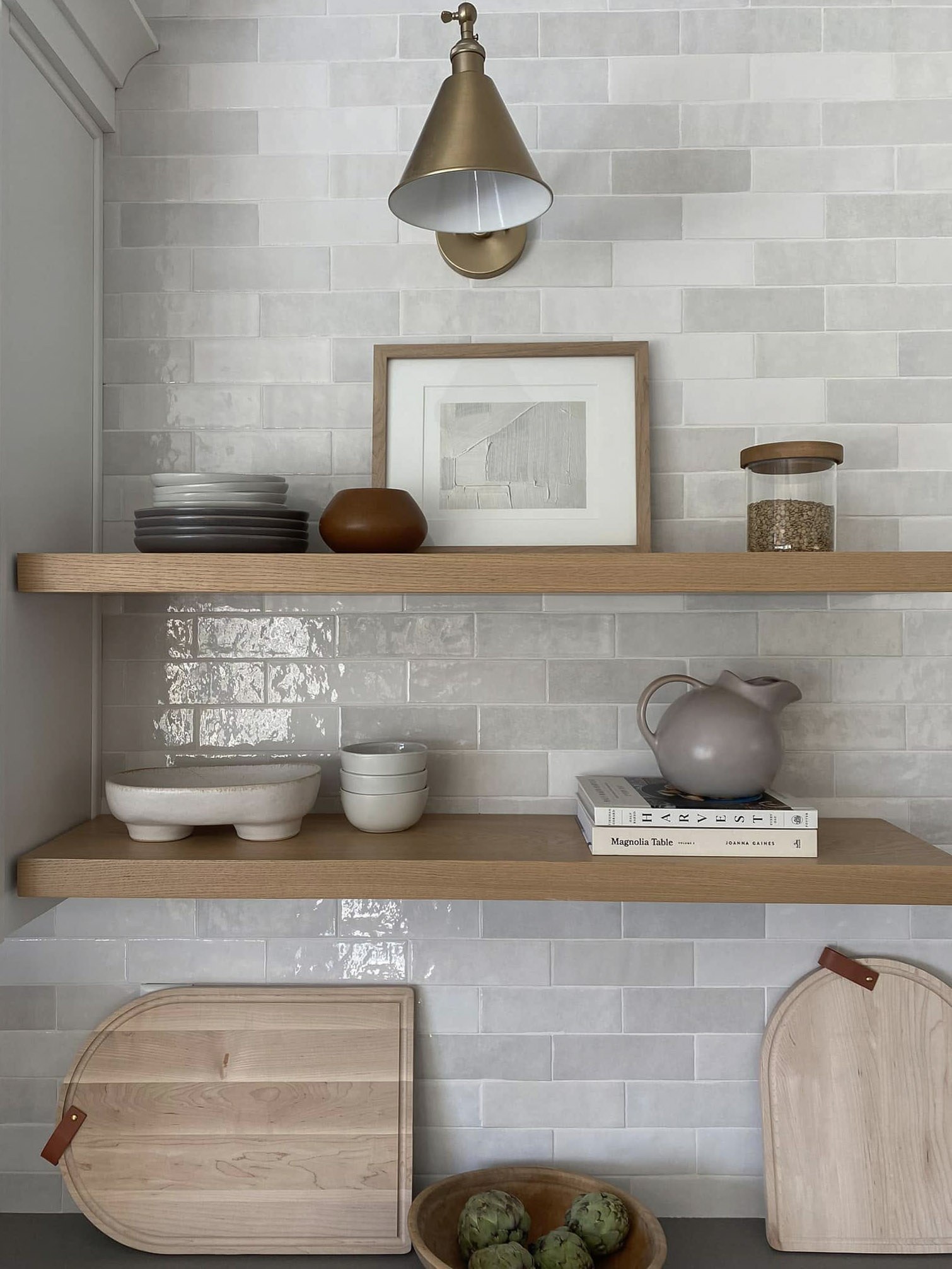 Oak floating shelves against glossy tile, holding stacked plates, ceramic bowls, a pitcher, and wooden cutting boards