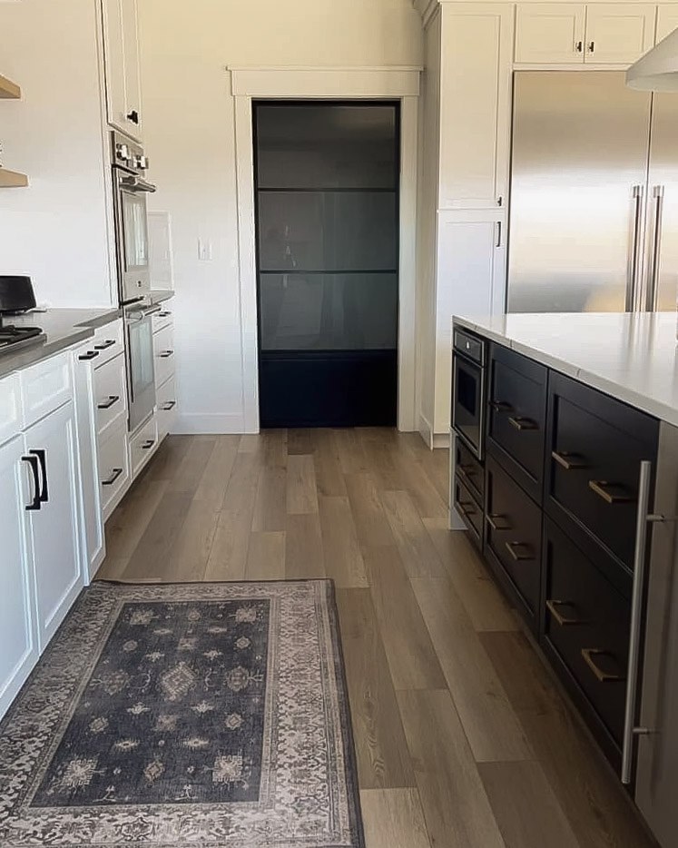 A galley view past the black island toward a dark glass pantry door, with a patterned runner on wide-plank floors
