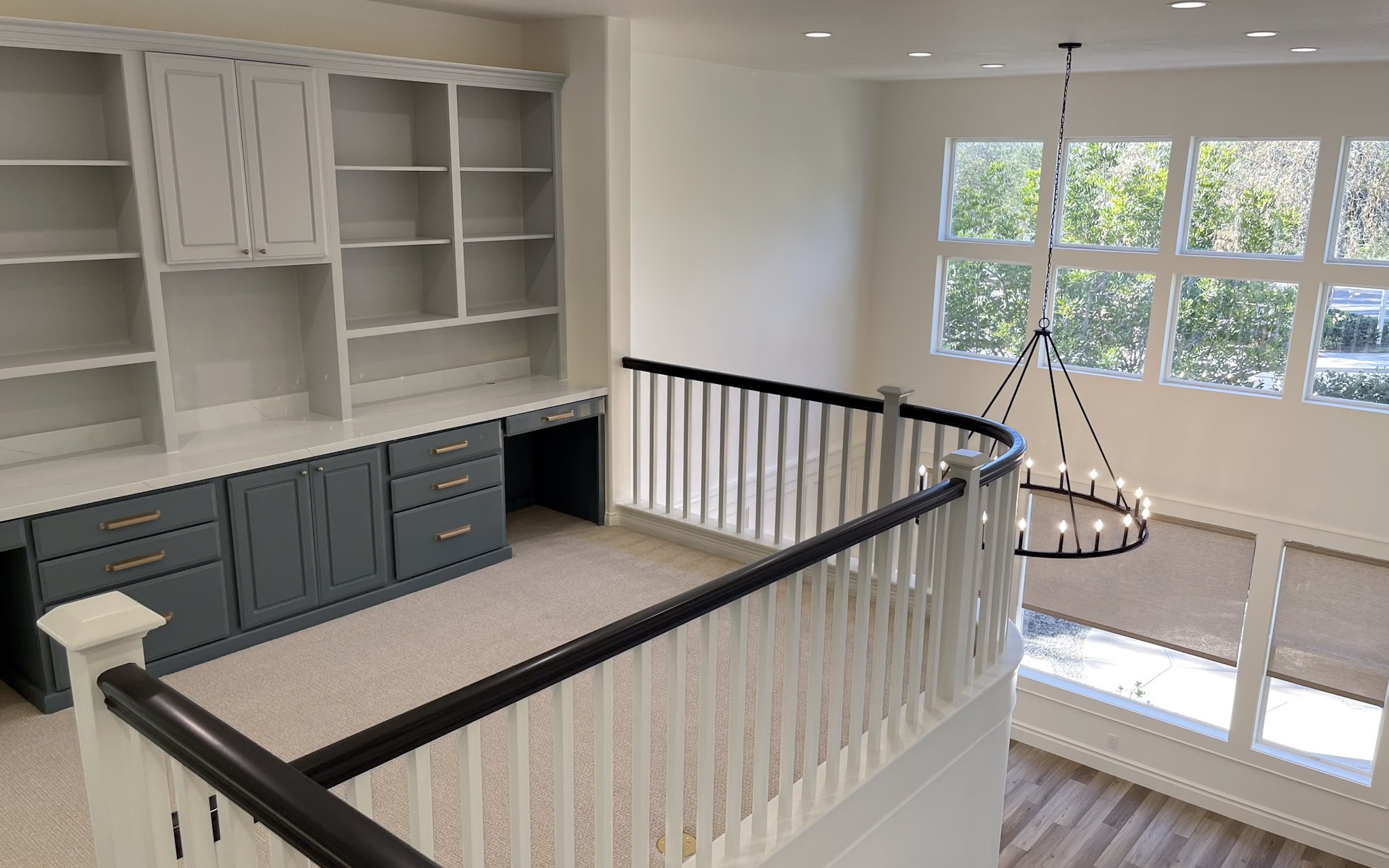 An upstairs landing overlooking a bright two-story room, with built-in shelving, a painted desk, and an iron ring chandelier
