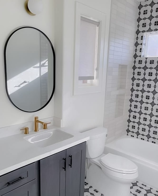 A bathroom with a gray vanity, brass faucet, black oval mirror, and black and white patterned tile around the tub