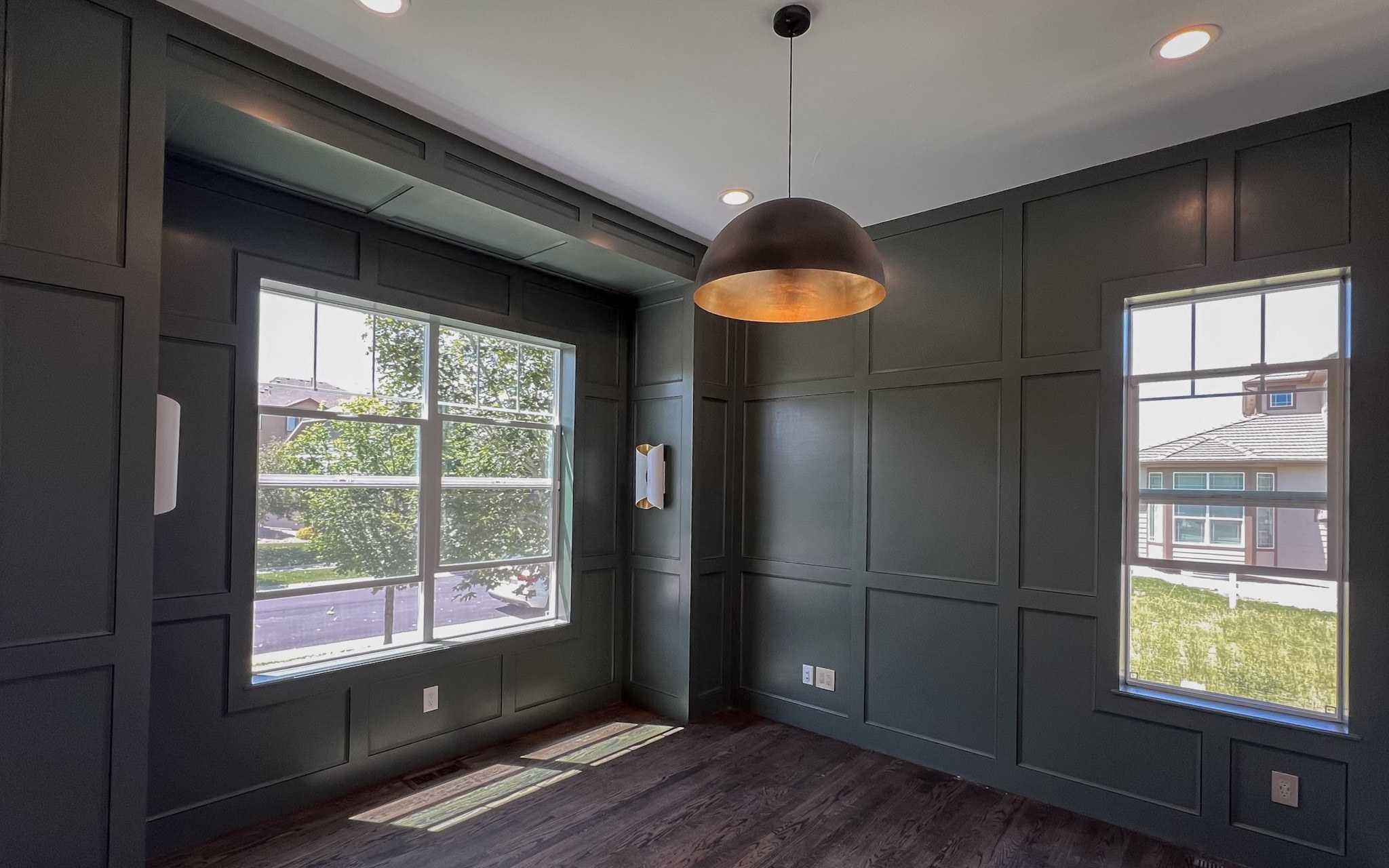 A corner room in deep green board-and-batten paneling, with a bronze dome pendant and daylight through two windows
