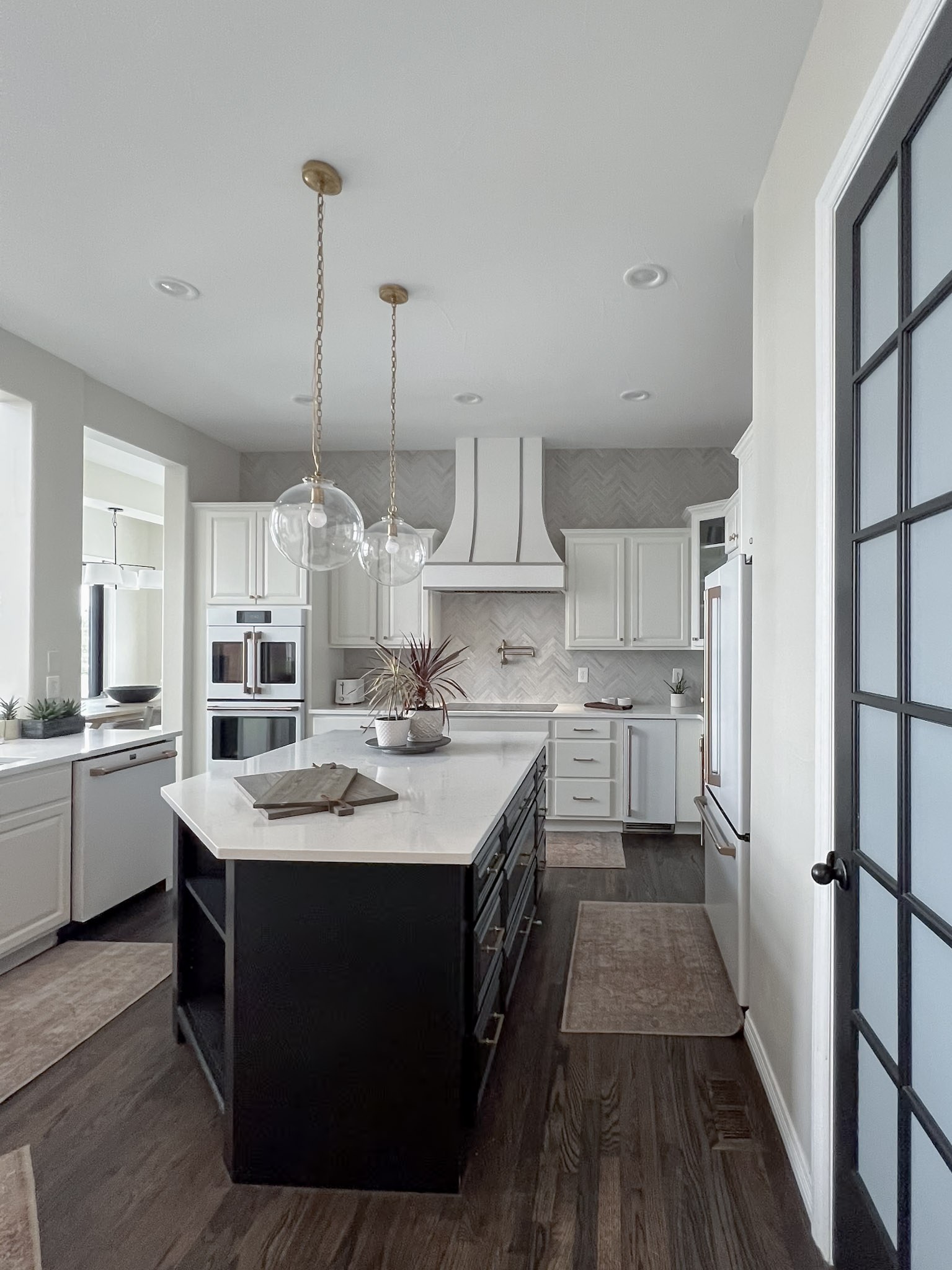 A white kitchen with a black island, brass chain pendants, herringbone tile backsplash, and dark wood floors